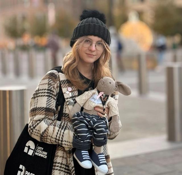 A person with long blonde hair wearing a black knit beanie, glasses, and a long plaid coat stands outdoors in a city setting. They are smiling and holding a stuffed animal rabbit dressed in a striped outfit and sneakers. The background of the image features tall buildings, bollards, and blurred autumn trees.