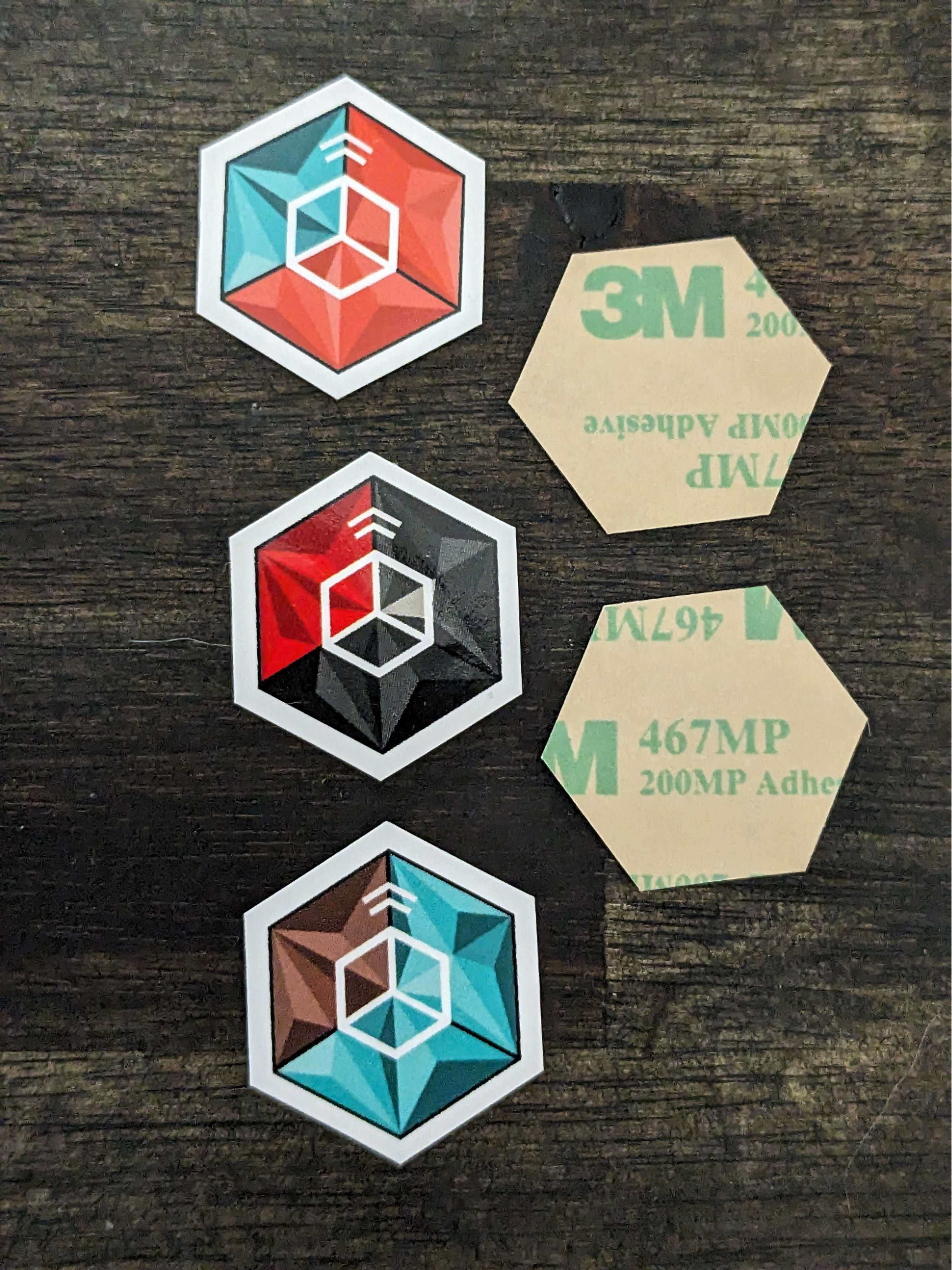 Hexagonal NFC stickers, on a wooden table, three with their blue/red backgrounds and cuboid icon designs facing up, two with 3M adhesive showing.