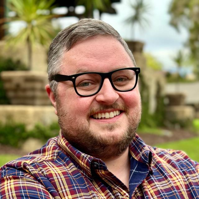 A smiling man with short gray hair and a beard, wearing black-rimmed glasses and a red, blue, and yellow plaid shirt. He is sitting outdoors in a lush garden with stone features and palm trees in the background.