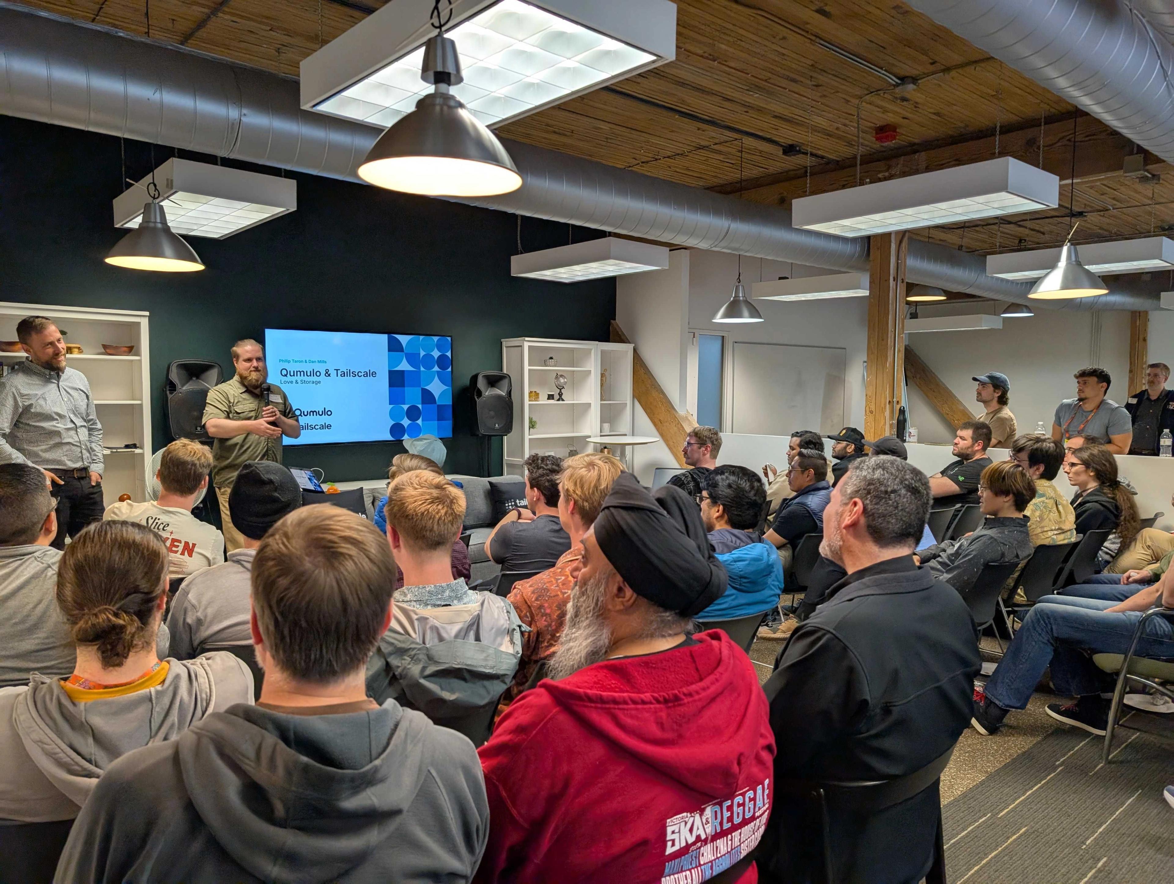 A group of approximately 30 people sitting together at the Seattle Tailscale Meetup, watching a presentation by Philip and Dan. There is a large presentation display that reads 'Qumulo and Tailscale, Love and Storage'. Most attendees are casually dressed. The background includes high ceilings with exposed beams, large hanging light fixtures and white walls.