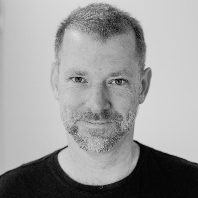 A man with a short well-groomed beard dressed in a white shirt, facing the camera with a calm demeanor.