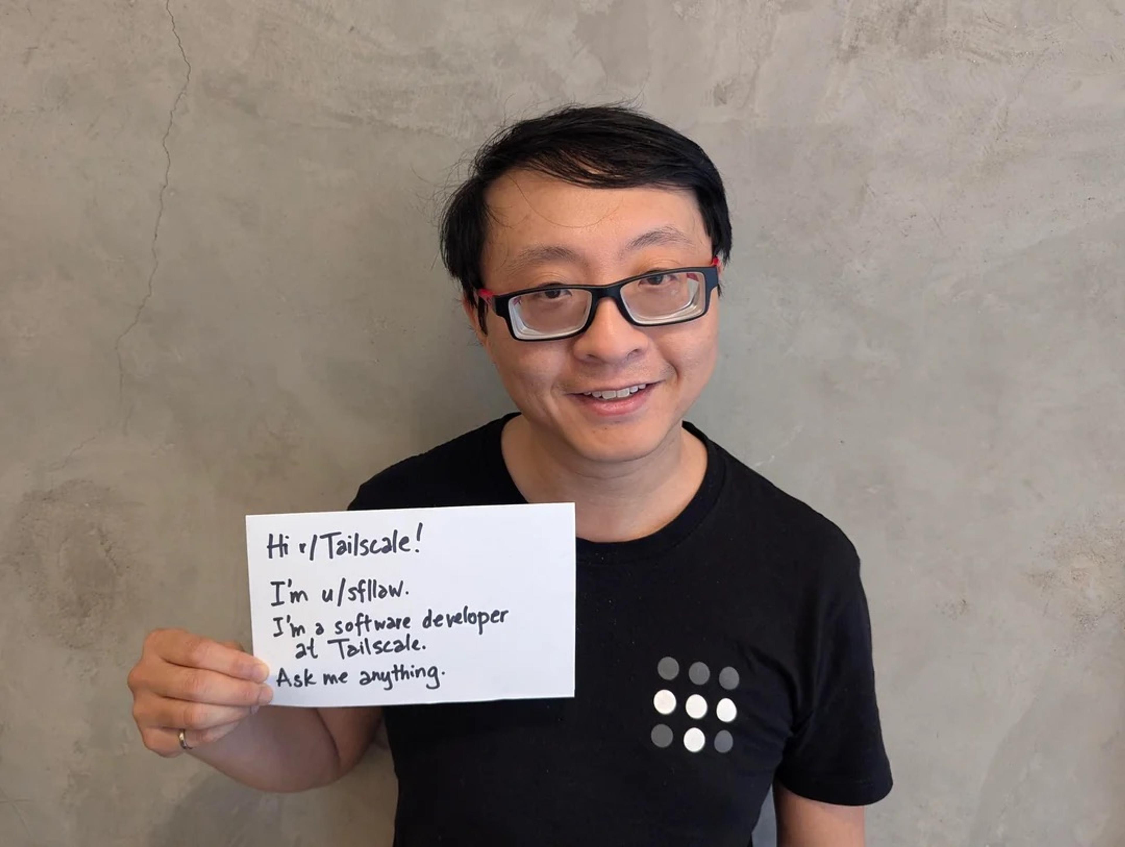 Simon Law, a Tailscale developer, wearing a black Tailscale loto T-shirt, holding up a notecard that reads "Hi r/Tailscale! I'm u/sflaw. I'm a software developer at Tailscale. Ask me anything."