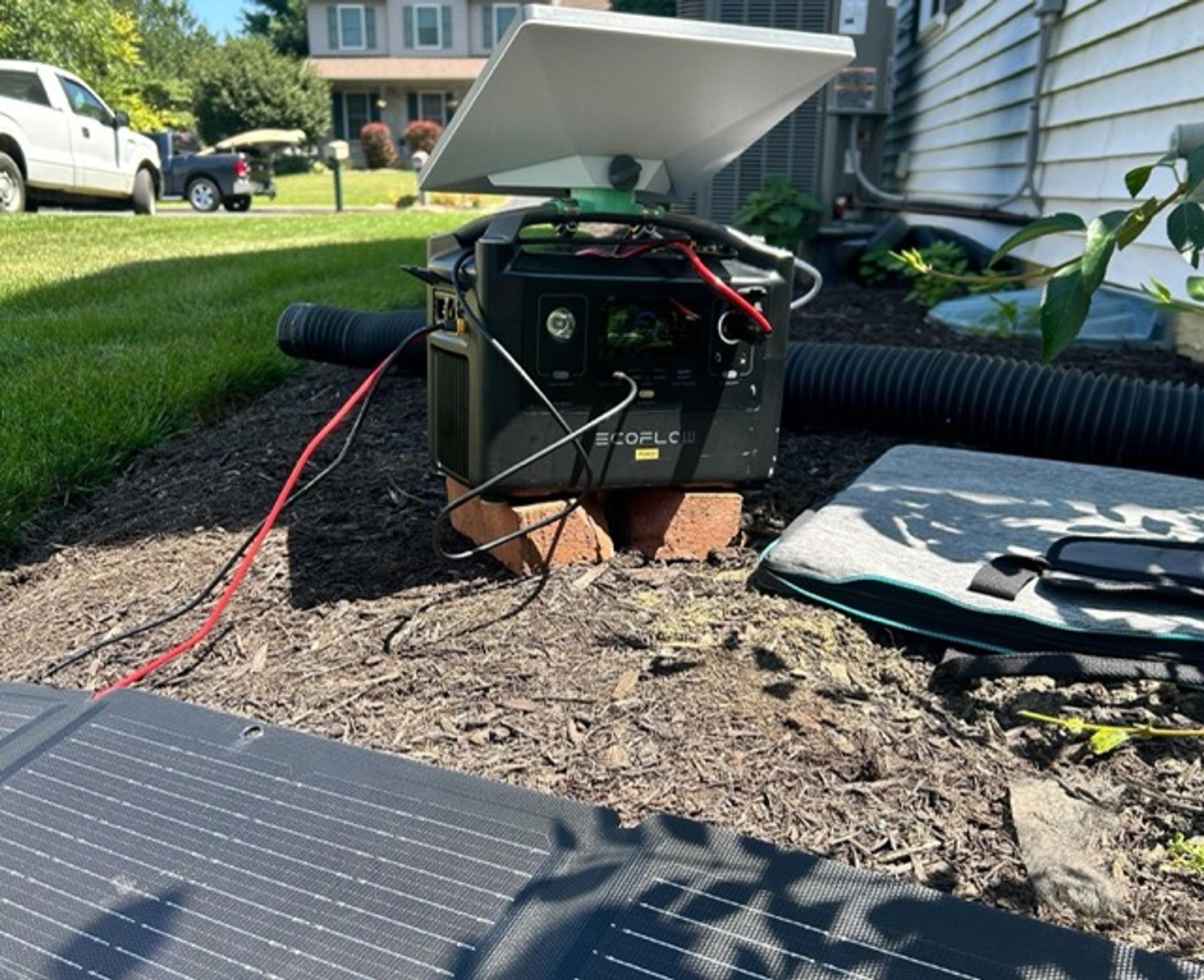 An EcoFlow Pro battery, with a satellite internet dish mounted on top through a small green plastic mounting receptacle, with wires leading from the stacked devices to a solar panel on the ground in the foregound.