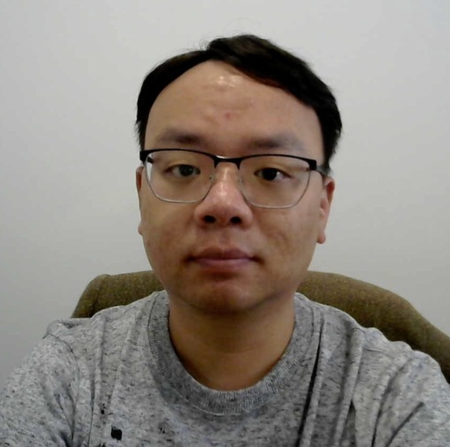 Man with dark hair and glasses, wearing a grey t-shirt in front of a white background.