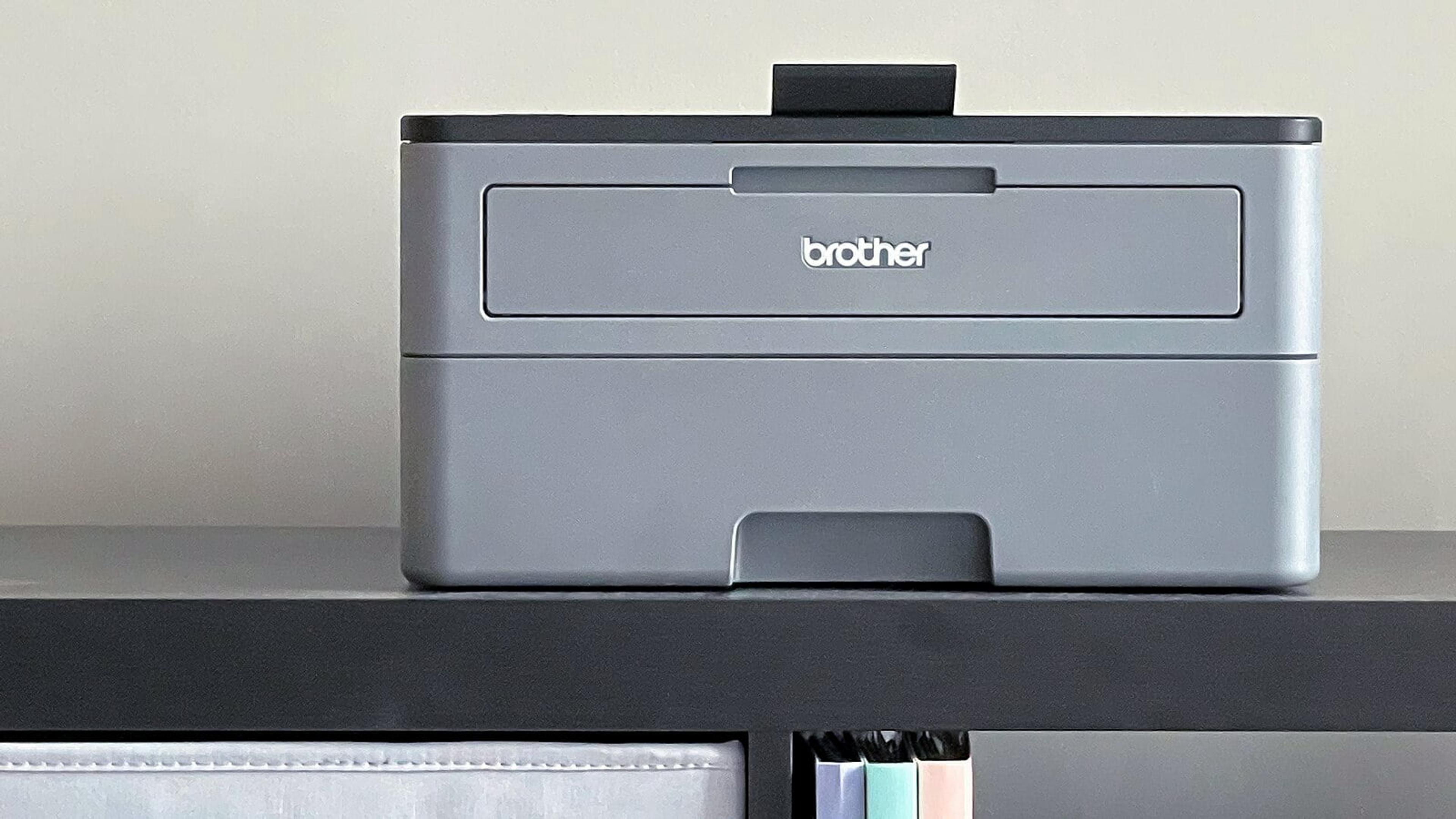 A gray, boxy laser printer, with a "brother" type logo embossed on the front, sitting on a black shelf with multi-colored binders and a fabric bin on lower shelves.