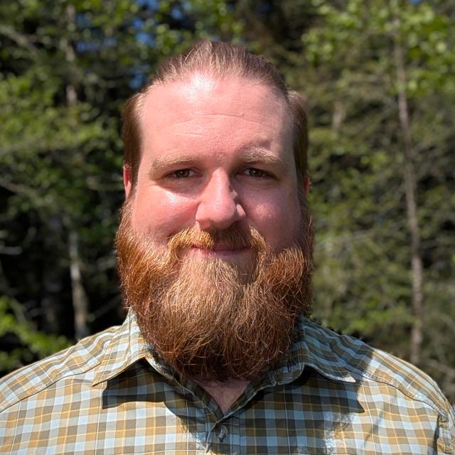 A man with a full reddish-brown beard and slicked-back hair, wearing a green and brown plaid shirt, smiling outdoors in a sunlit forest setting.