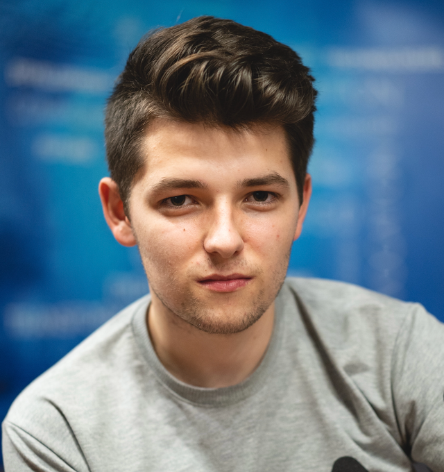 A man with short dark hair wearing a light gray shirt looks directly at the camera, with a blue blurred background behind him.