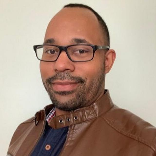 A man with short hair and a trimmed beard, wearing black rectangular glasses and a brown leather jacket over a dark blue shirt, smiling slightly in front of a plain light-colored background.