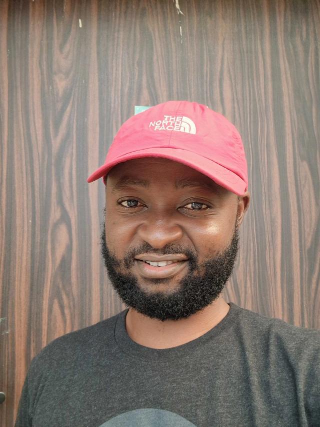 A smiling man wearing a red The North Face baseball cap and a dark gray T-shirt, with a short full beard. He is standing in front of a brown wood-grain wall, facing the camera in a close-up portrait.