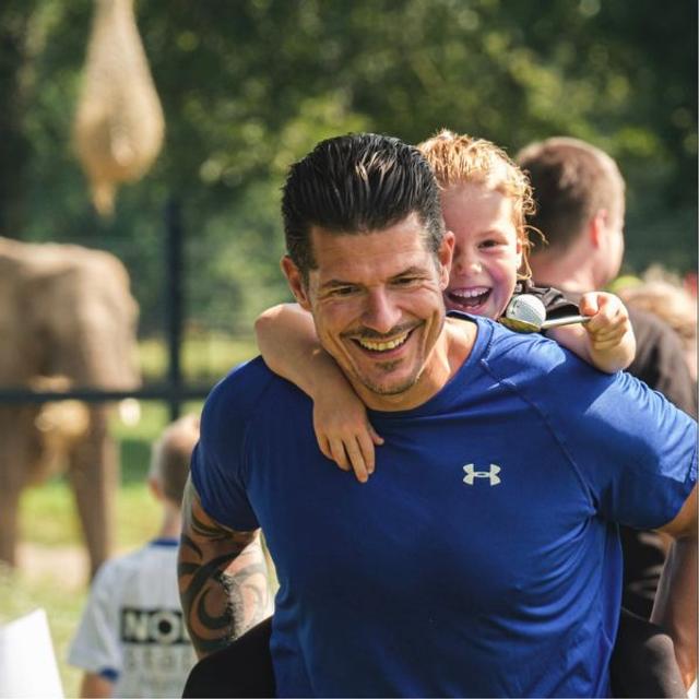 A man with dark hair wearing a blue tshirt and carrying a child on his back.