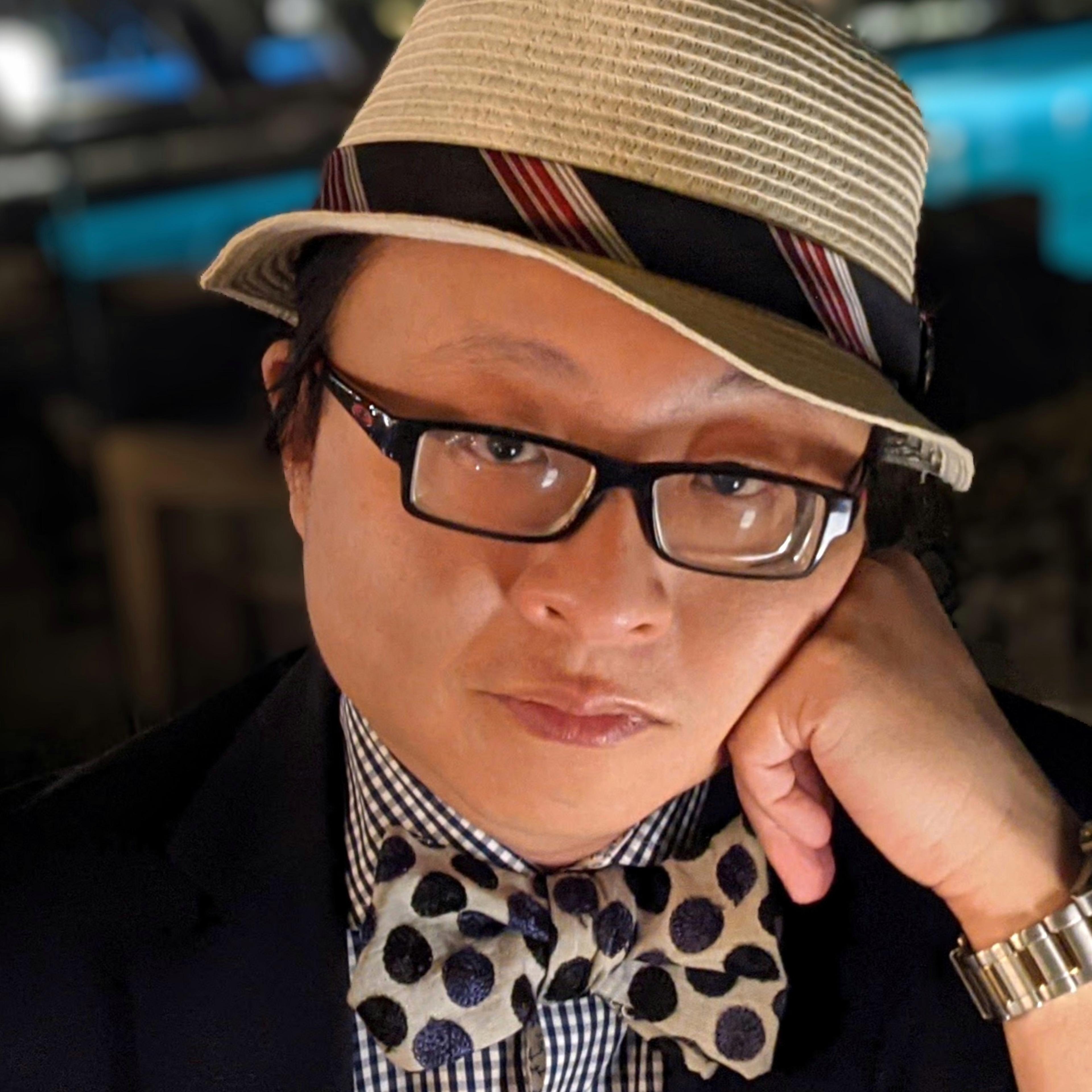 Headshot of Simon Law, wearing a beige hat, black-rimmed glasses, and a polka dot bow tie. 