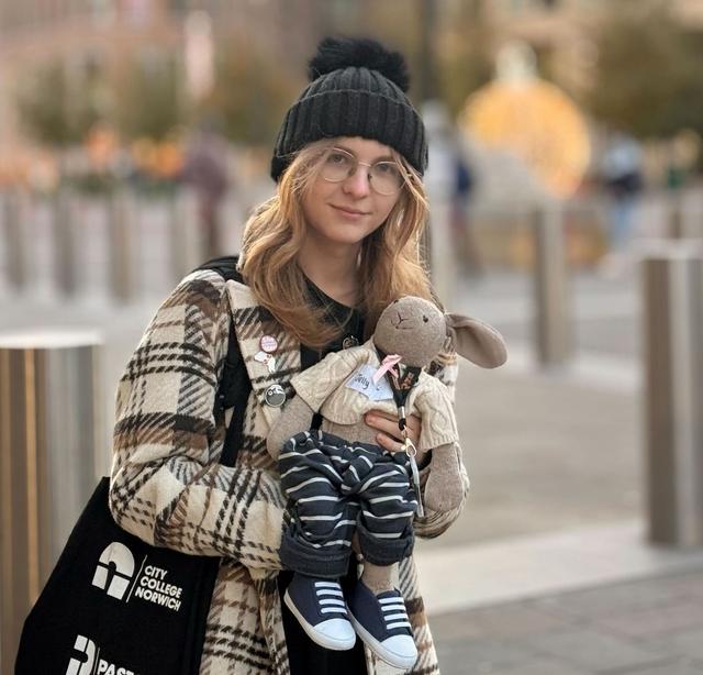 A person with long blonde hair wearing a black knit beanie, glasses, and a long plaid coat stands outdoors in a city setting. They are smiling and holding a stuffed animal rabbit dressed in a striped outfit and sneakers. The background of the image features tall buildings, bollards, and blurred autumn trees.