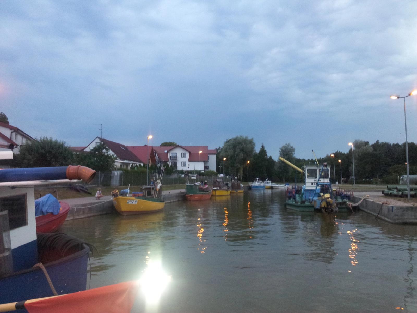 Basen portowy w Rowach o zmierzchu, kolorowe łodzie rybackie przy obu nabrzeżach i światła latarni odbijające się w wodzie.