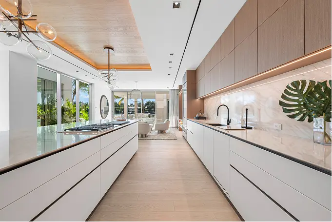 Long, light-filled kitchen with modern lighting, wood flooring, and white and wood cabinetry.