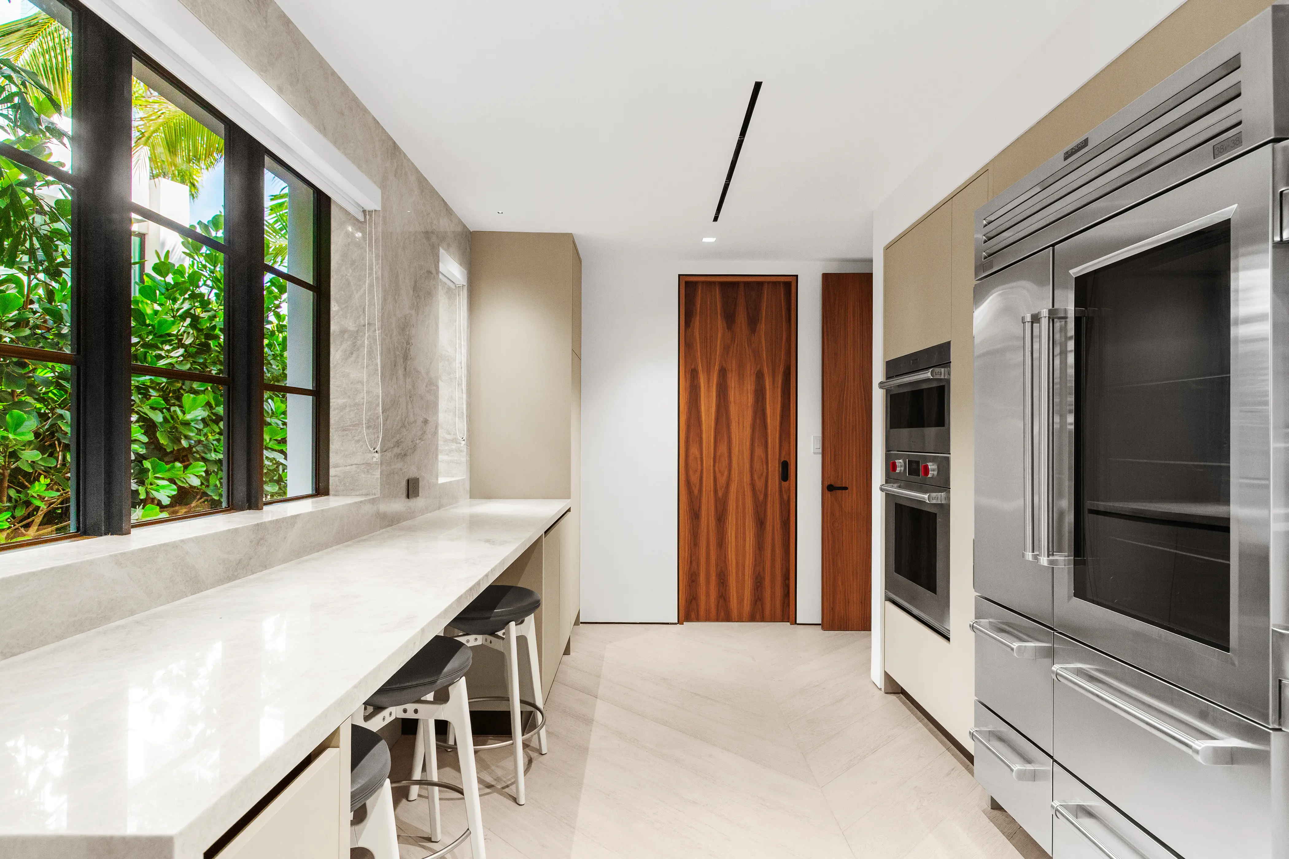 Luxury Miami Beach kitchen featuring marble countertops, custom cabinetry, professional-grade appliances, and expansive black-framed windows overlooking tropical landscaping.