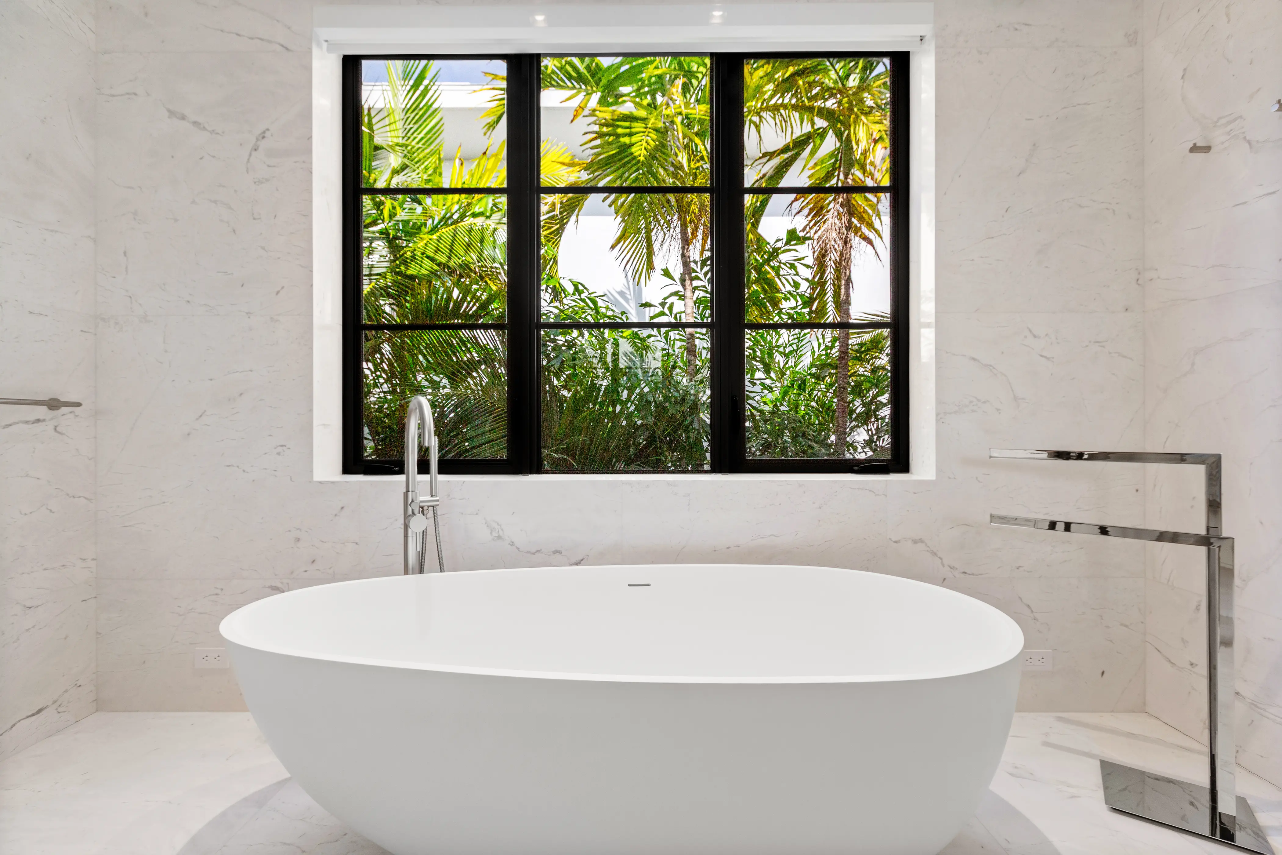 Luxury Miami Beach bathroom featuring a freestanding soaking tub, floor-to-ceiling marble surfaces, black-framed windows, and refined contemporary fixtures.