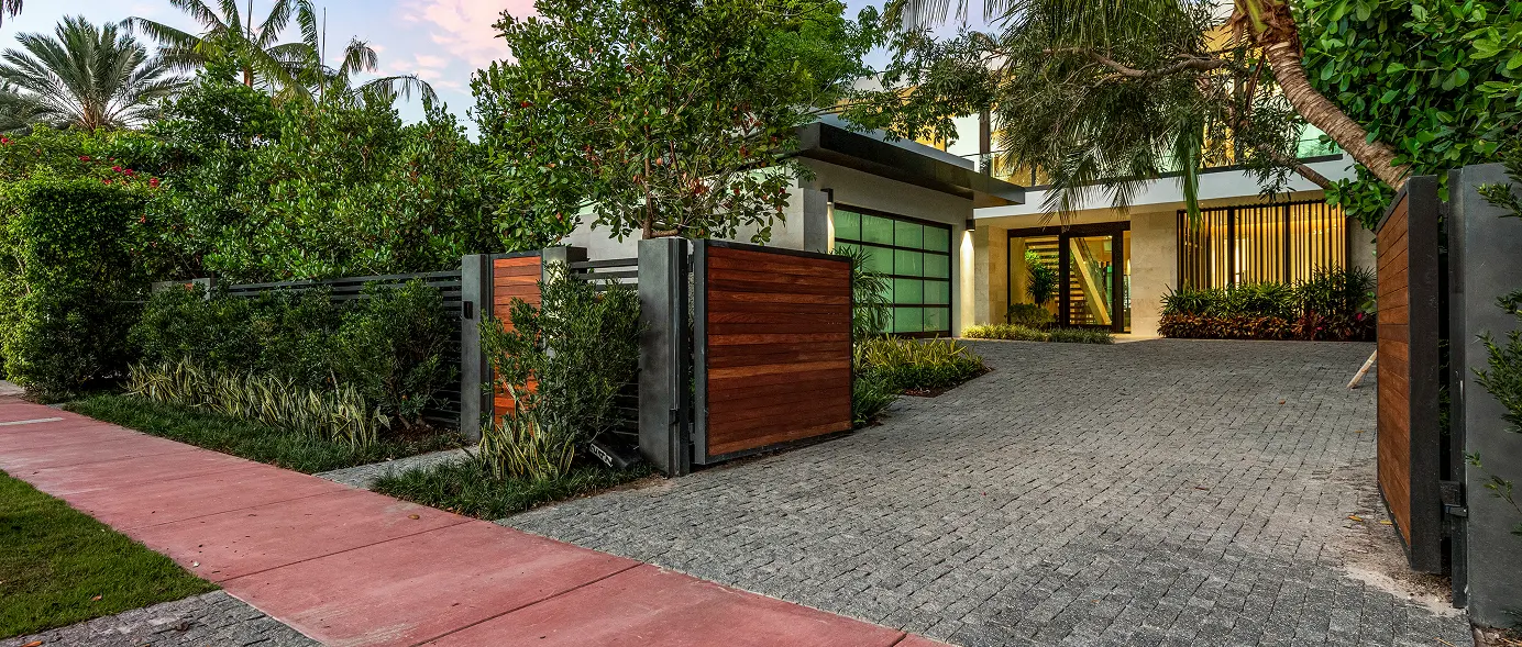 Approach view of the home from the stone driveway, past the glass garage doors and mahogany gate.