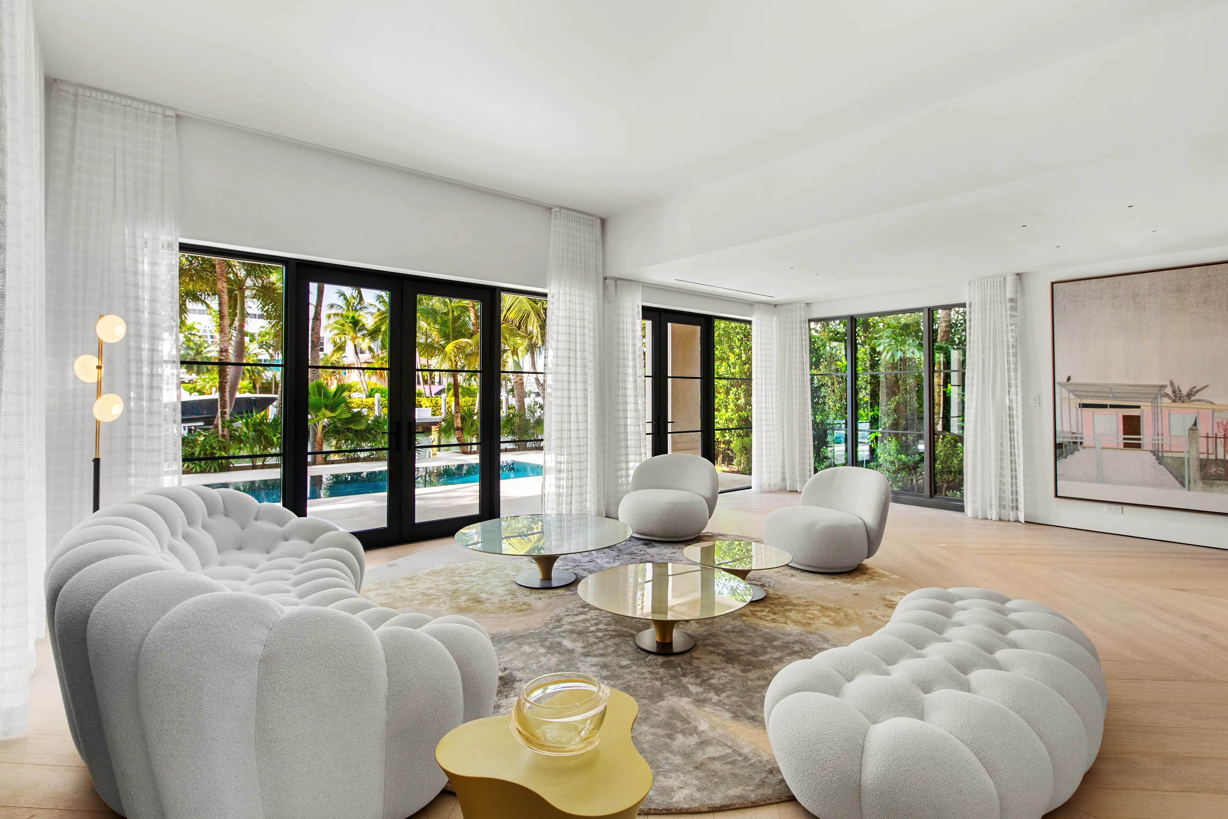 Bright luxury living room featuring floor-to-ceiling windows, custom furnishings, neutral finishes, and views of the tropical pool area.