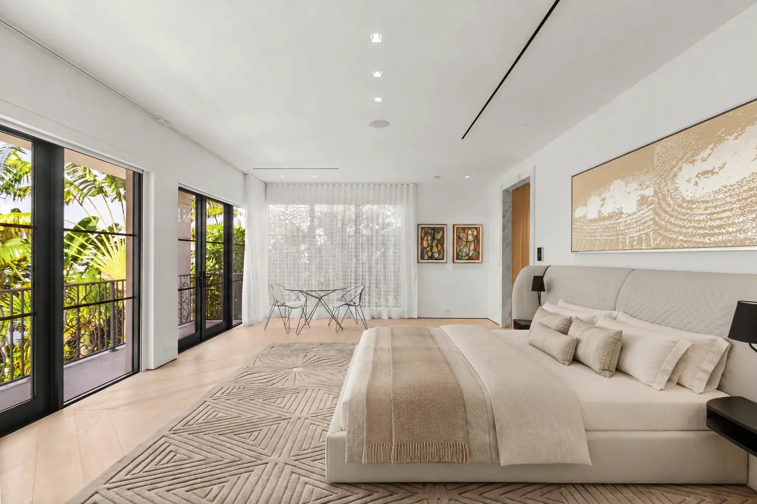 Light-filled primary bedroom featuring soft neutral finishes, contemporary furnishings, recessed lighting, and expansive black-framed doors opening to tropical views.