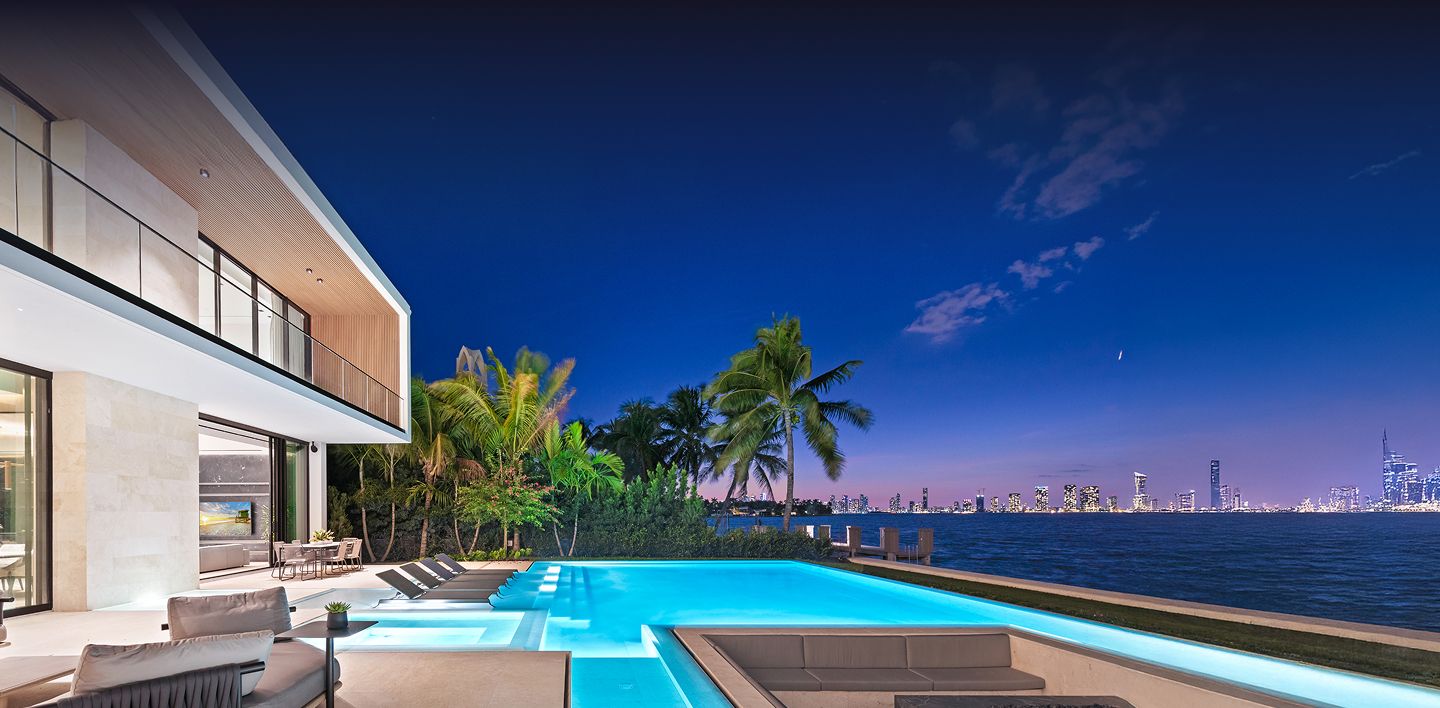 Modern waterfront residence at twilight with a glowing infinity pool, palm trees, and panoramic Miami skyline views across the bay