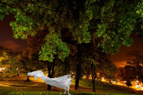 Dame med hvitt flagrende tekstil rundt seg står i en kunstig belyst park. 