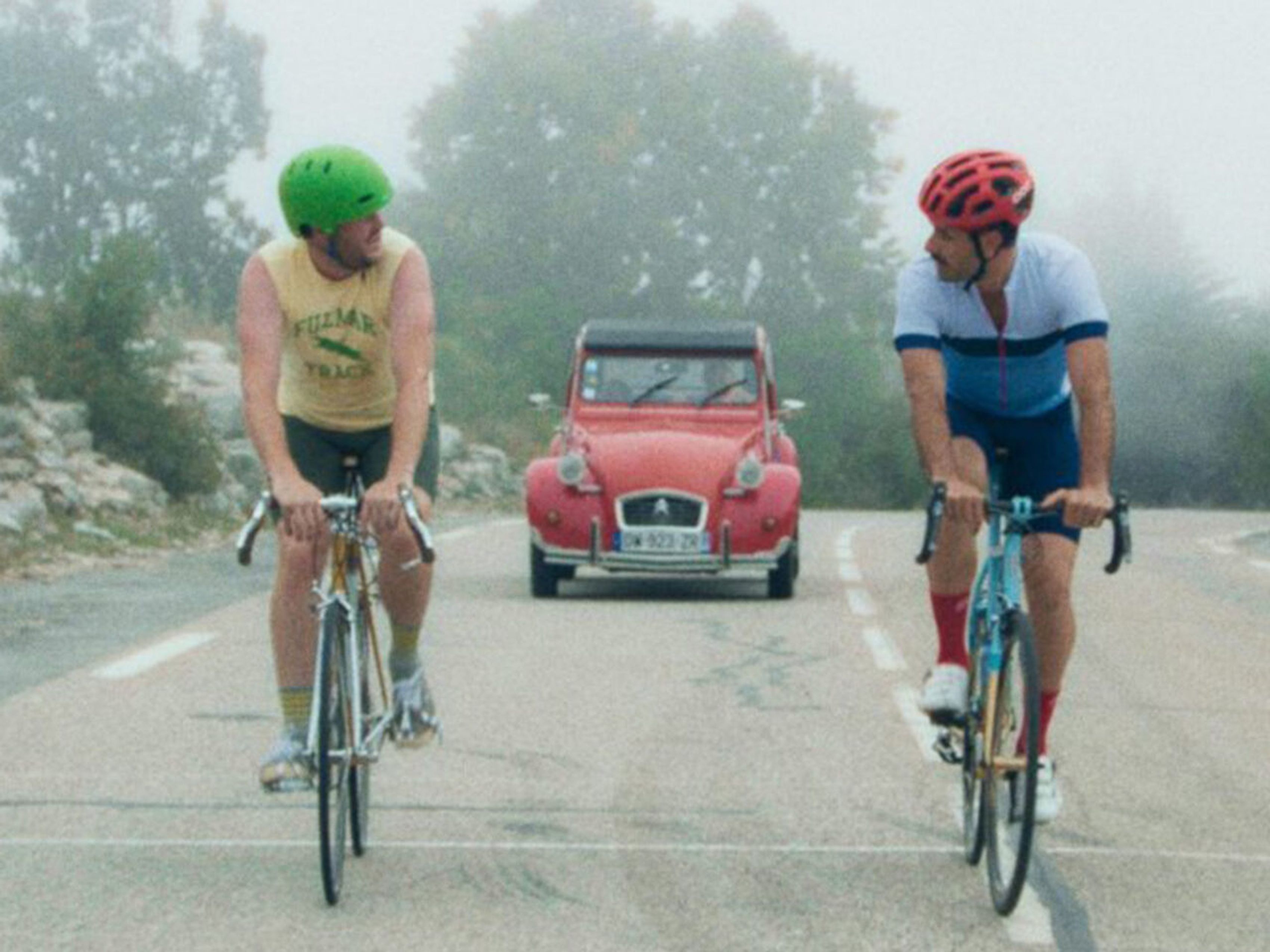 two men on bicycles talking with red car in background