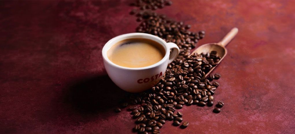 A Costa Coffee cup surrounded by roasted coffee beans and a wooden scoop filled with coffee beans on a dark surface.