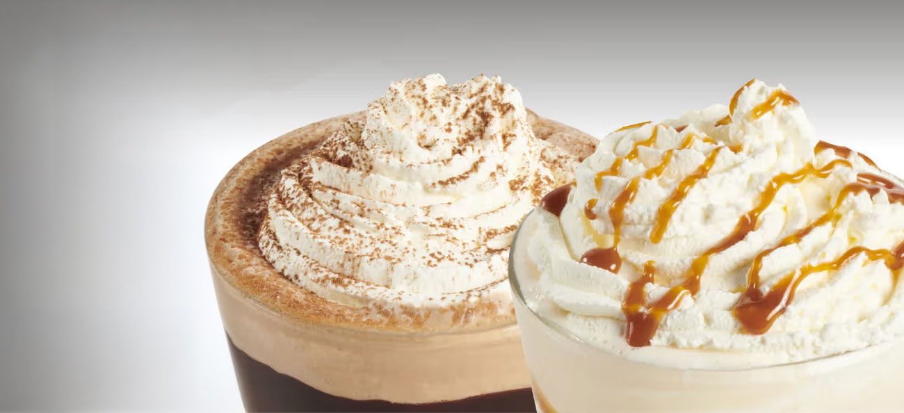 Picture of two iced coffee drinks topped with Cream. The left one is iced mocha frappe, topped with cream and chocolate powder, and the right one is a Salted Caramel Toffee Frappe, topped with cream and a drizzle of toffee sauce
