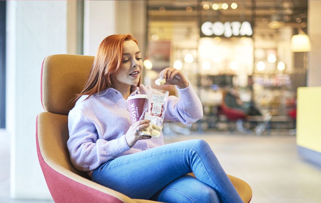 A customer enjoying a Costa Coffee beverage and snack while seated, with a Costa Coffee store visible in the background.