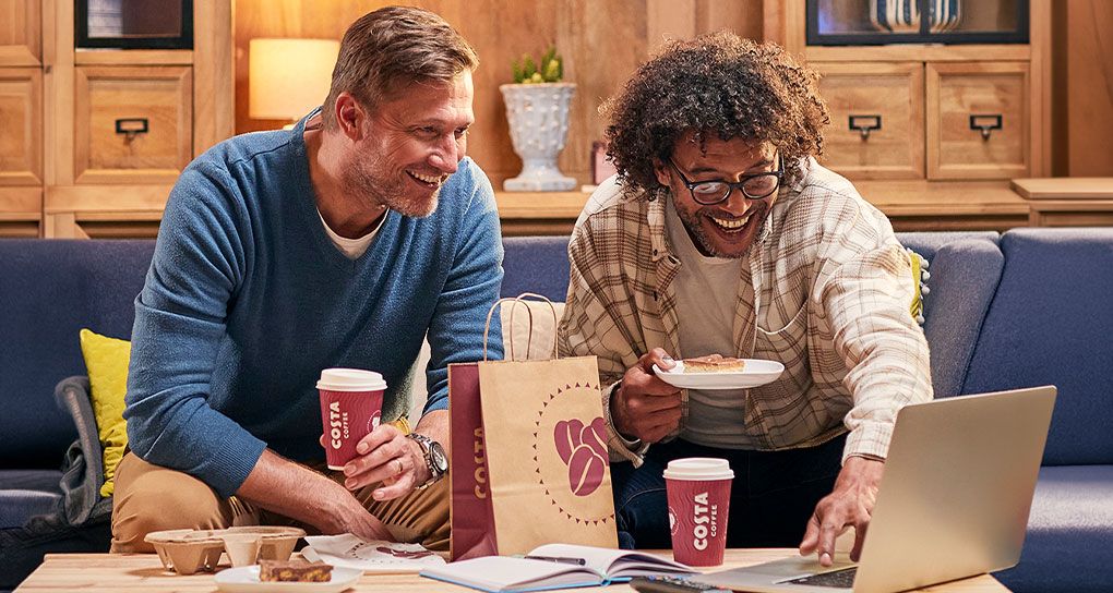 Two people enjoying Costa Coffee drinks and snacks while using a laptop.