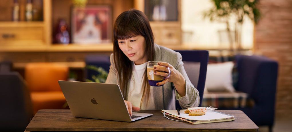 Person using a laptop while holding a coffee at a table in a Costa Coffee café, with a notebook and snack nearby.