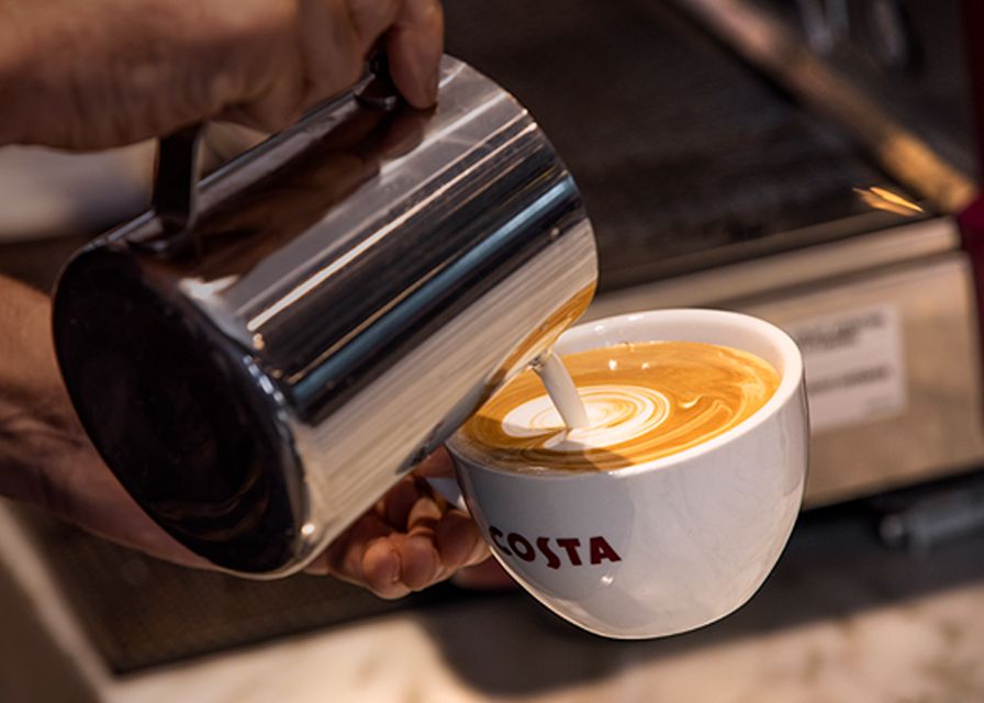 Barista vertiendo leche de una jarra para hacer arte latte en taza de Costa Coffee