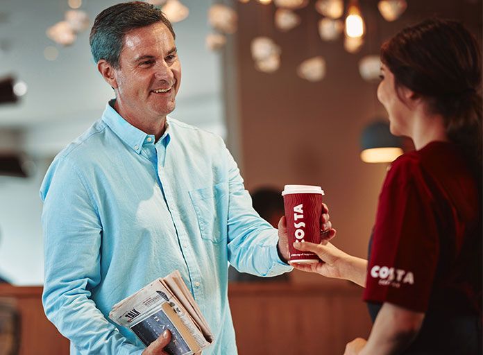Muž přebírá kávu Costa Coffee s sebou od člena týmu Costa Coffee v kavárně Costa Coffee.