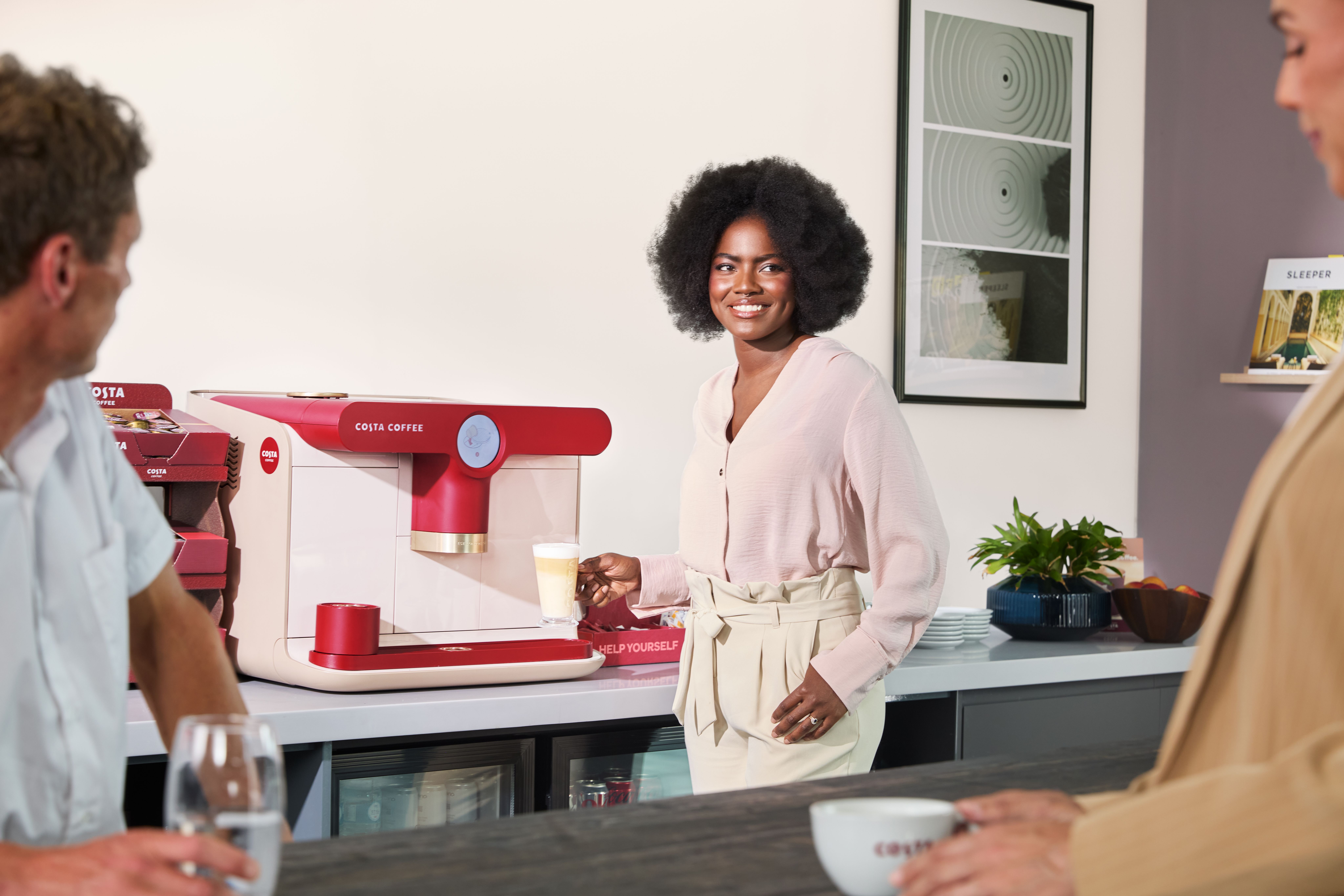Person dispensing a coffee drink from a Podio Costa Coffee machine in a workplace break area.