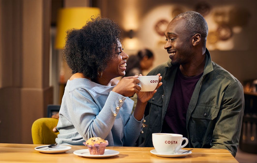 Two people sitting at a Costa Coffee café table, enjoying hot drinks and a muffin in a warm, modern coffee shop setting.