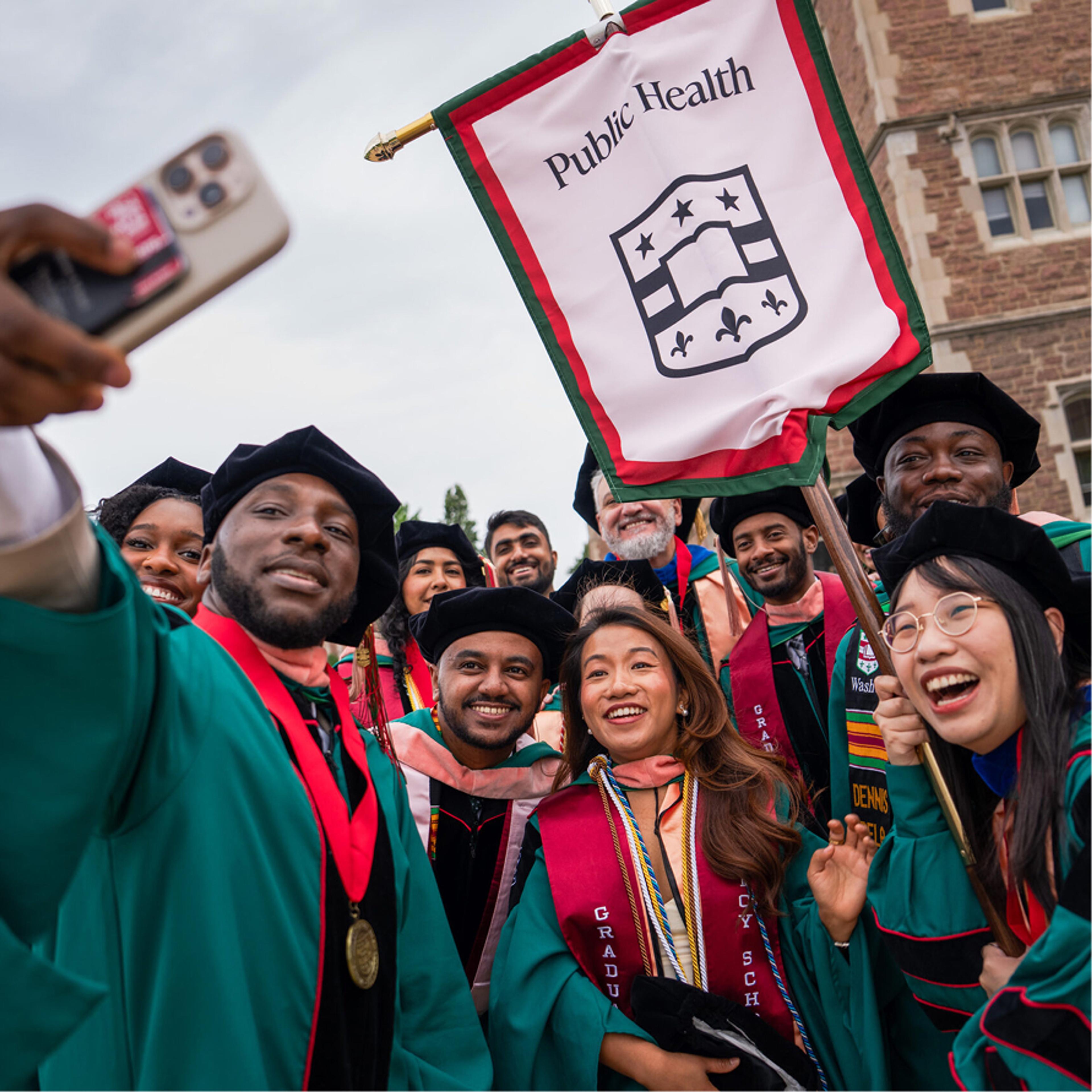 Graduates in green regalia taking a selfie under a Public Health banner