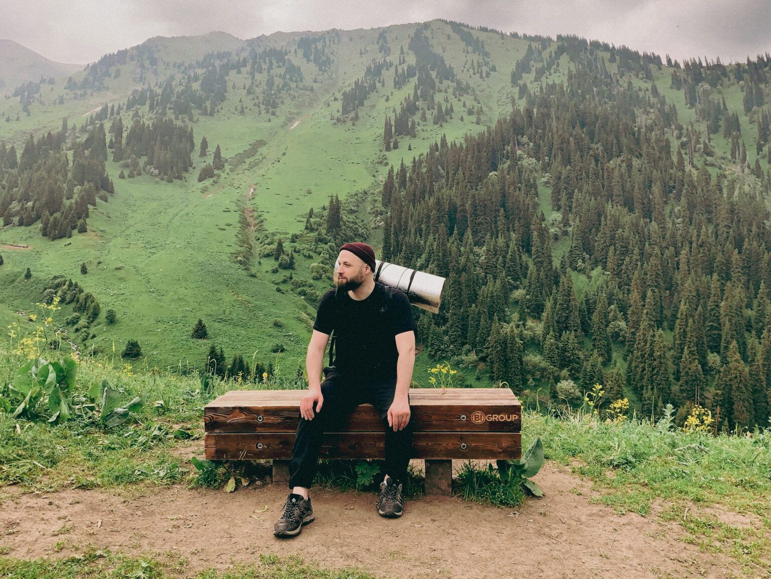 Hiker with a rolled sleeping mat sits on a bench overlooking lush, forested mountains.