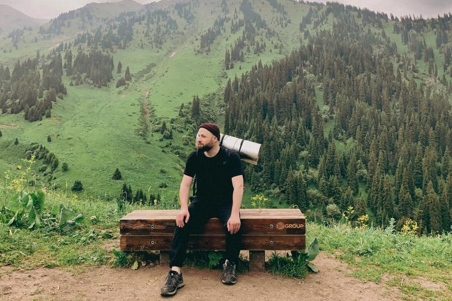 Hiker with a rolled sleeping mat sits on a bench overlooking lush, forested mountains.