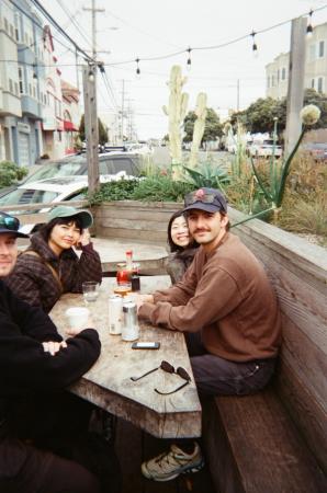 Four friends gather around an outdoor picnic table beside a cactus garden on a city street.
