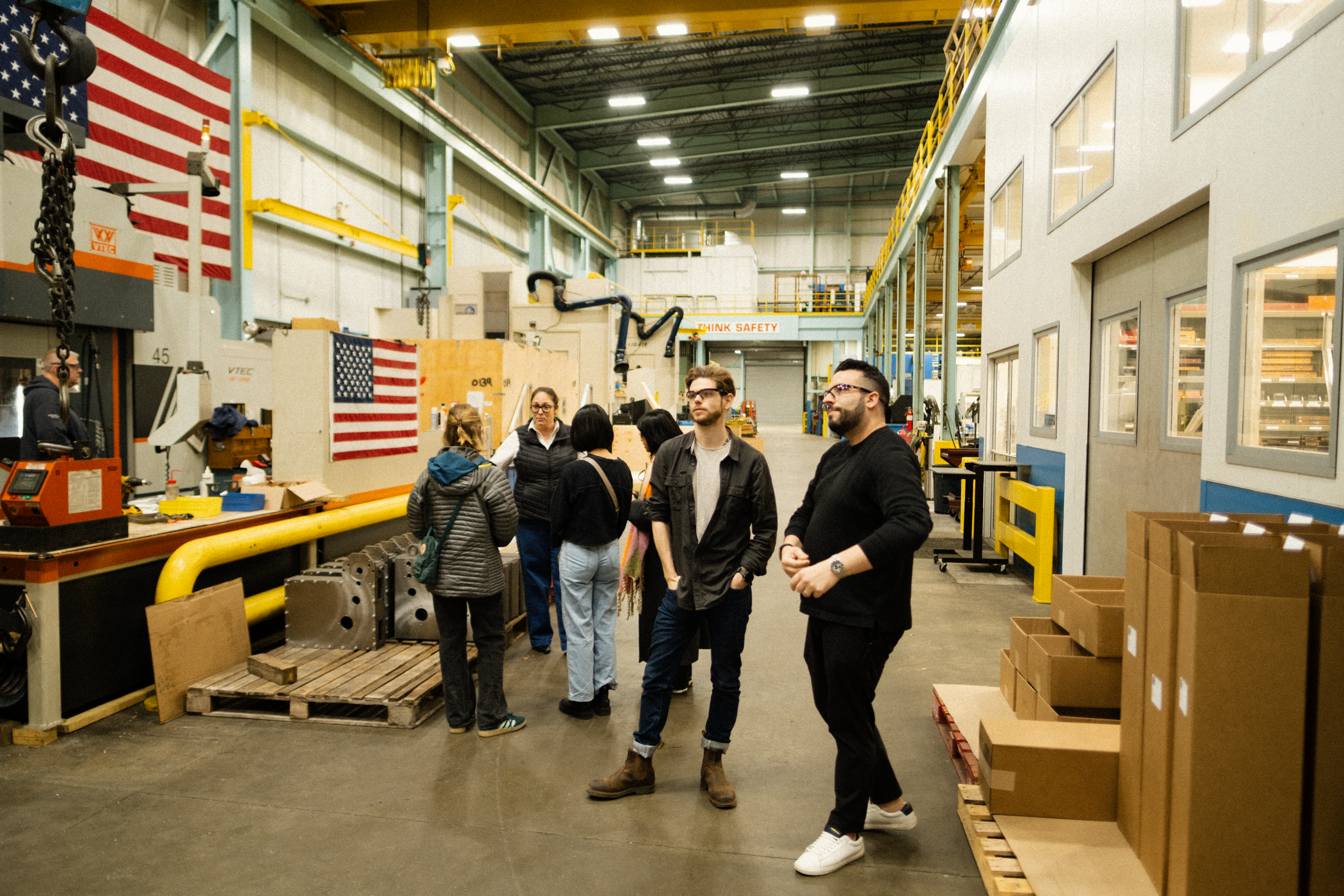 Temper And Forge team members tour a manufacturing facility with machinery, boxes and American flags.