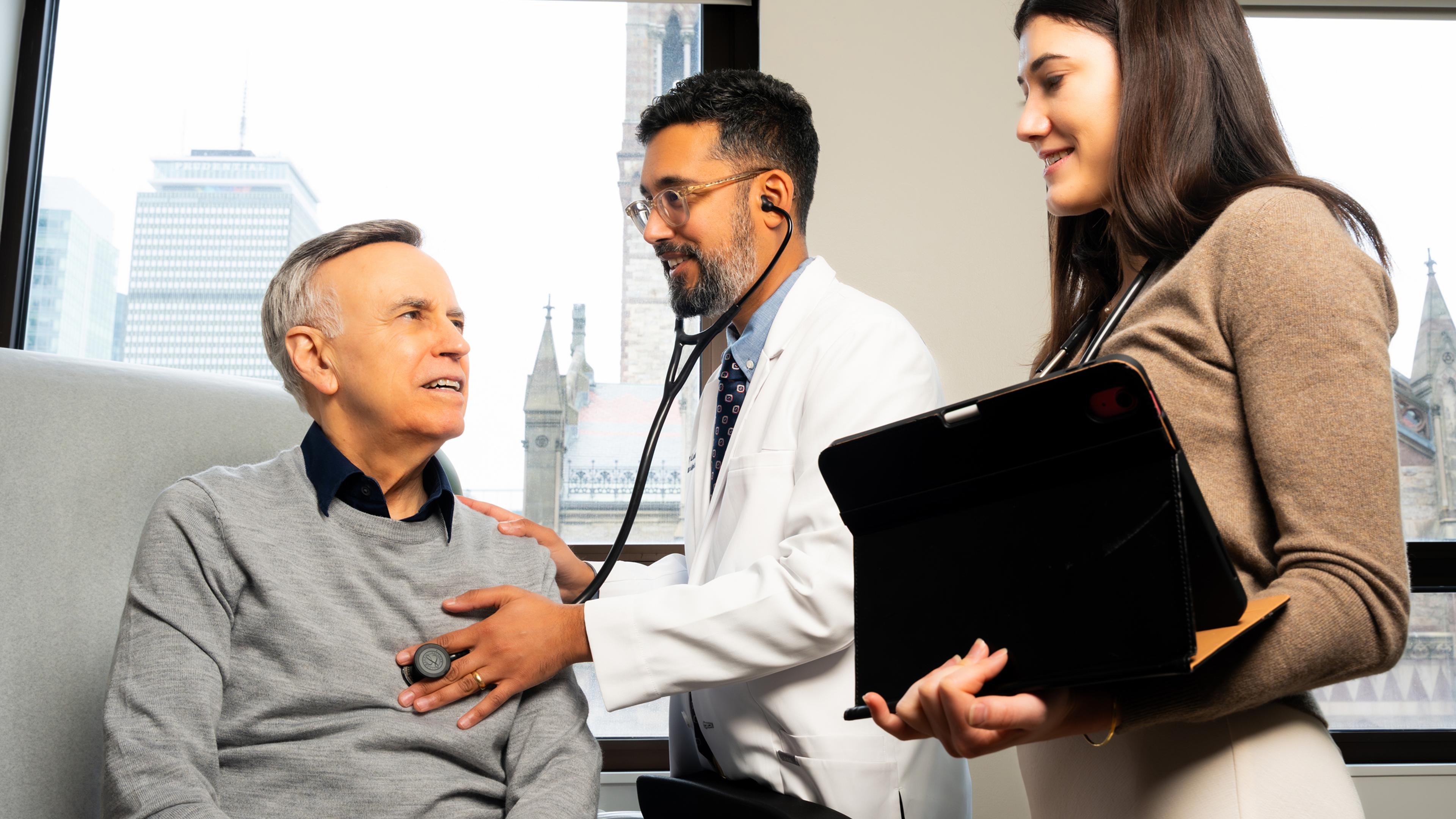 Doctor listening to a patient's heart with a stethoscope while a woman stands by holding a tablet computer