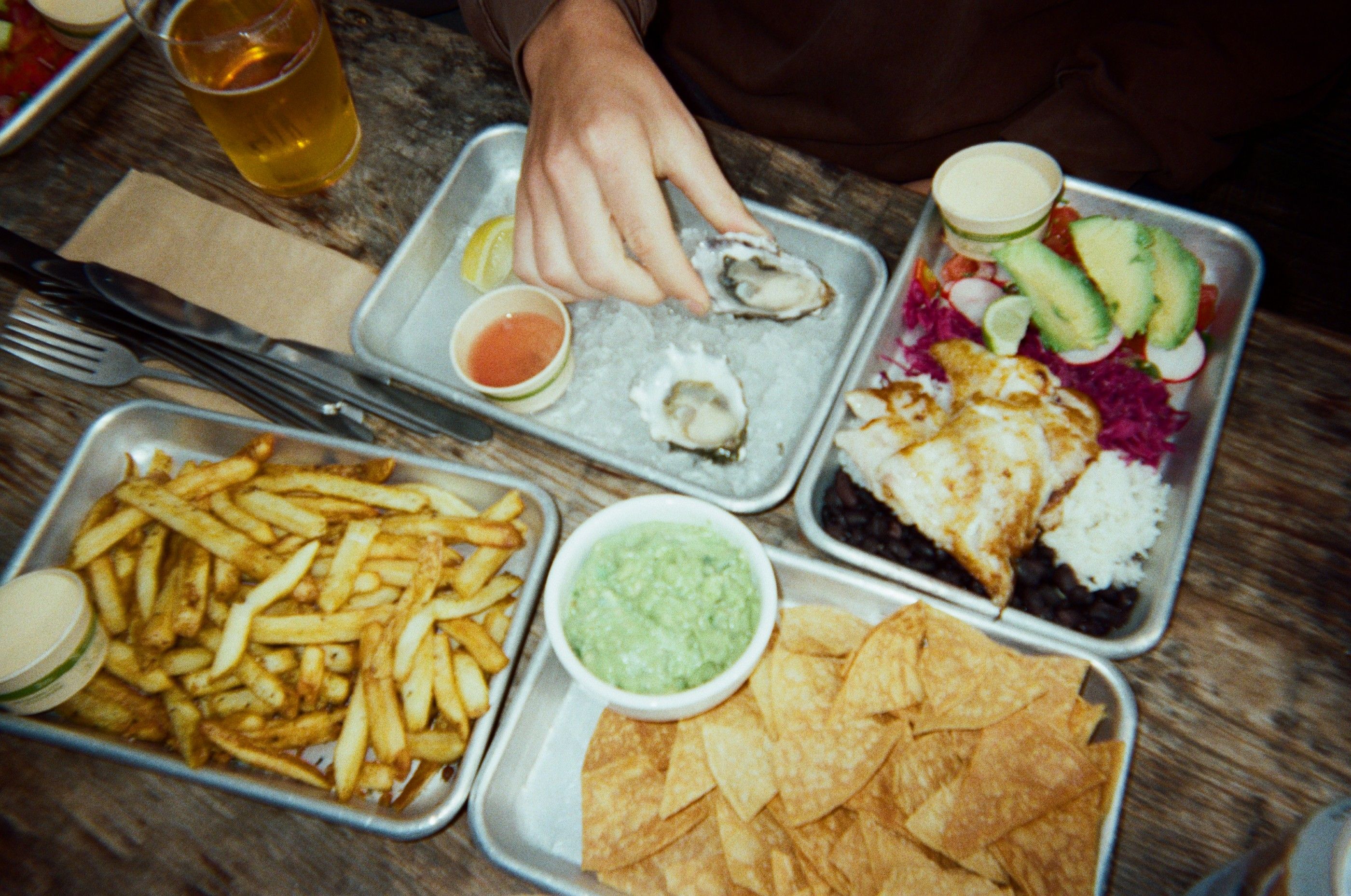 Diner reaches for an oyster beside trays of fries, chips and guacamole, and a rice bowl with avocado.
