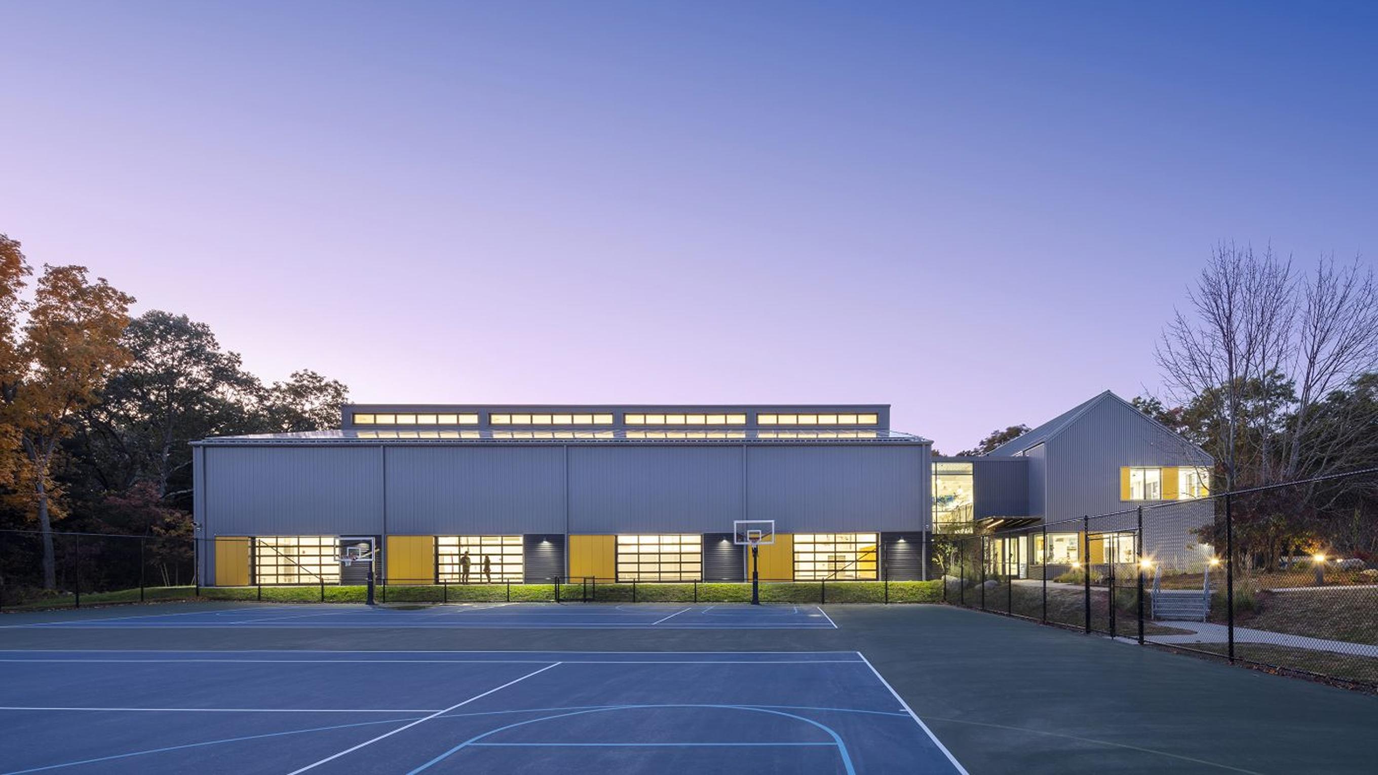Photo of a building at dusk. The lights are on inside and a basketball court is seen in the foreground