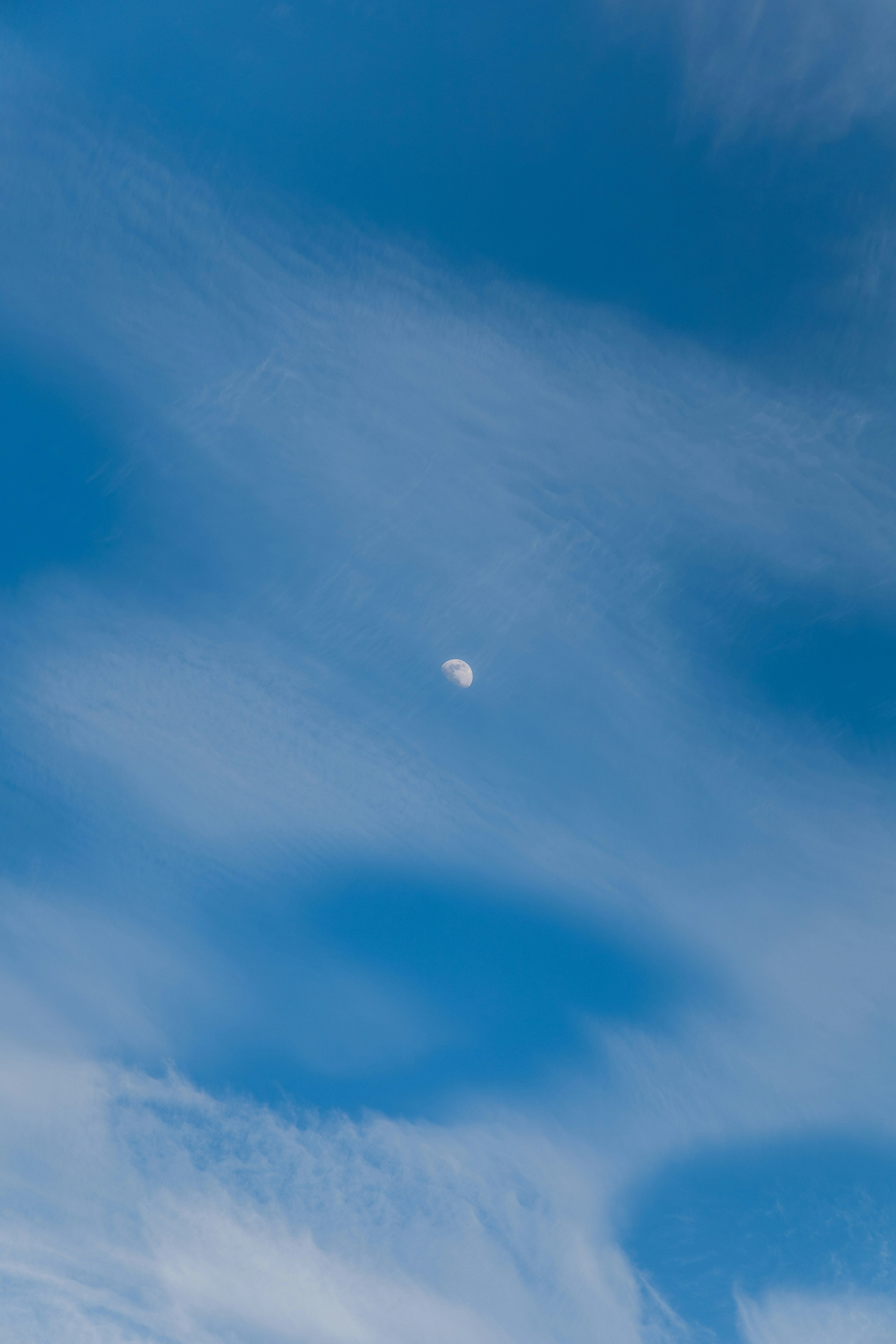 Photo of the daytime sky with the moon in the center an wispy cloud