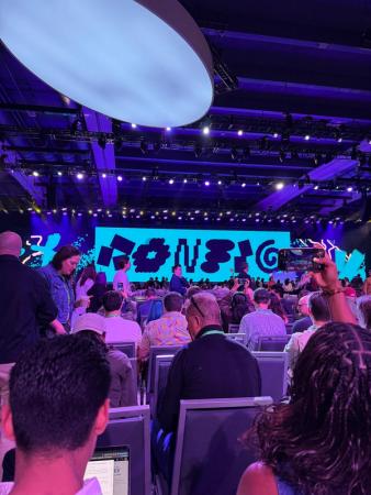Crowd seated in a purple-lit conference hall facing a stage with a colorful abstract display.