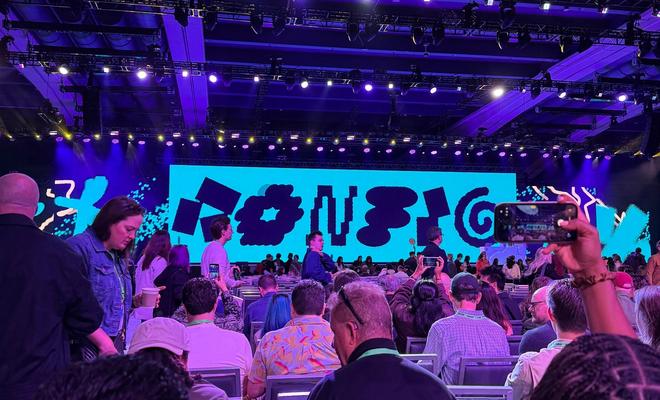 Crowd seated in a purple-lit conference hall facing a stage with a colorful abstract display.