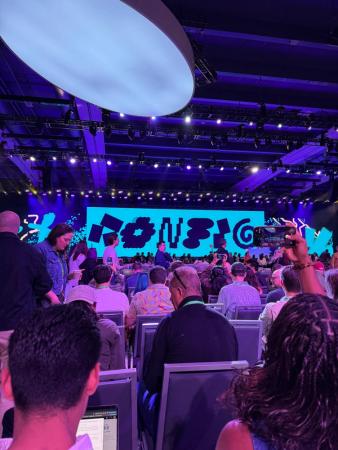 Crowd seated in a purple-lit conference hall facing a stage with a colorful abstract display.