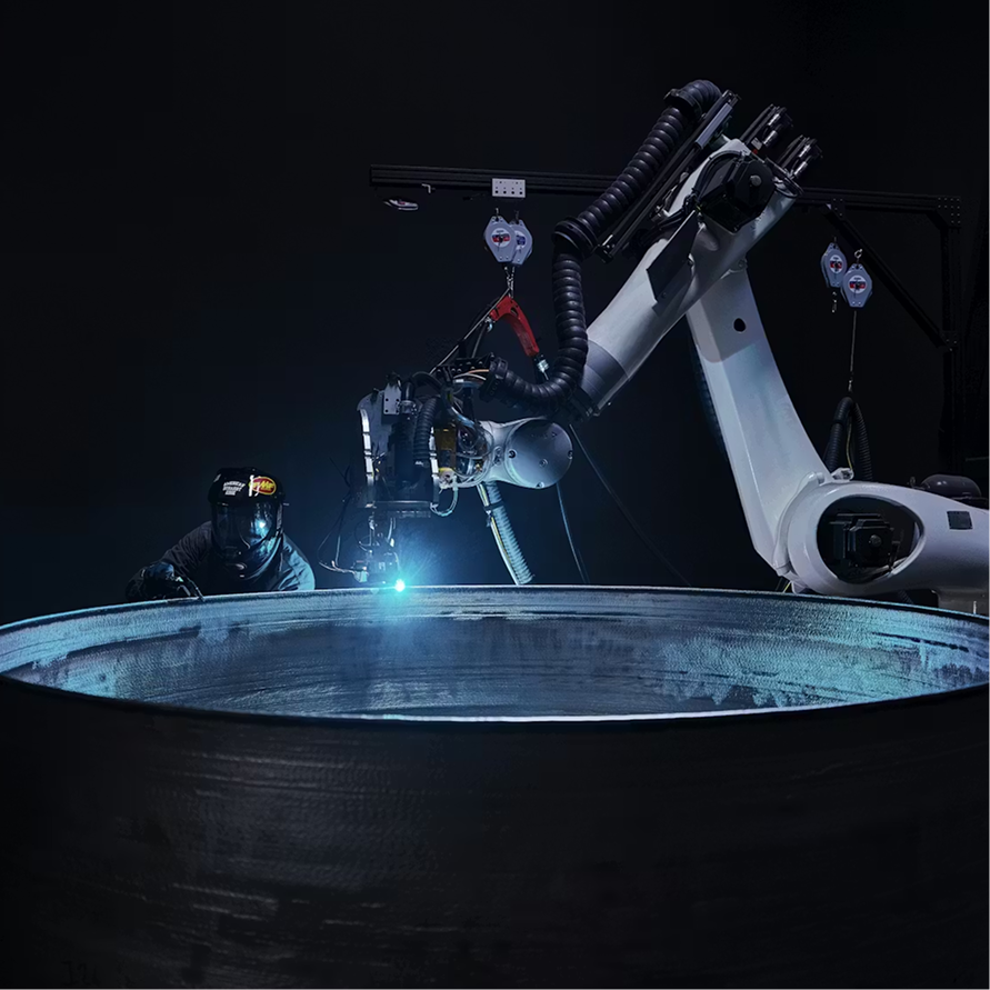 A robotic arm welding the rim of a large steel drum as a masked worker looks on