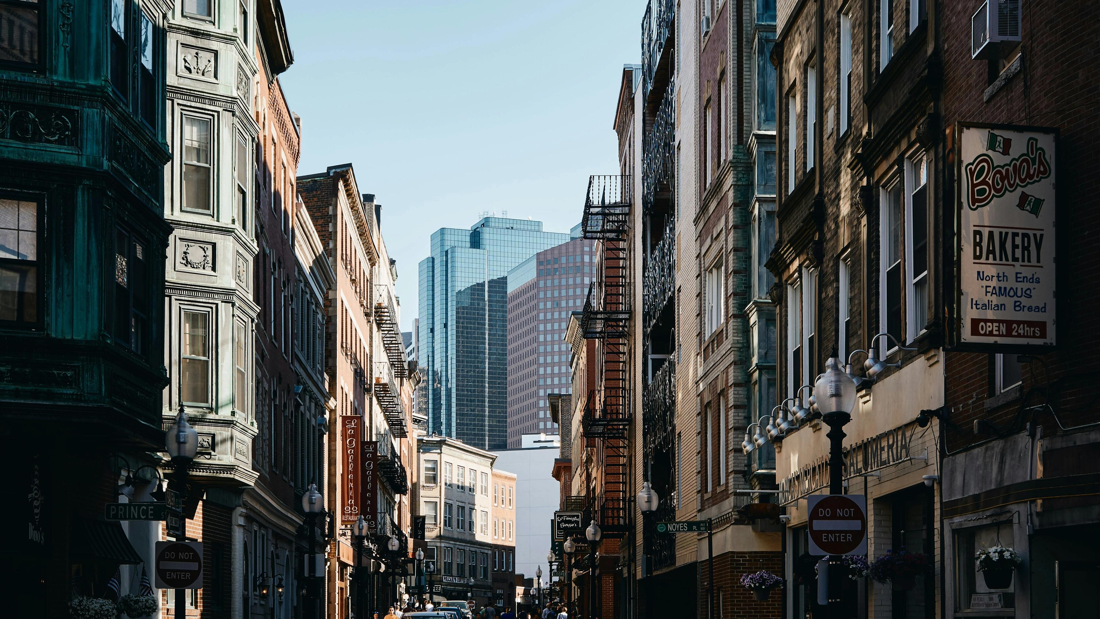 A narrow downtown Boston street of brick buildings, with Financial District towers beyond