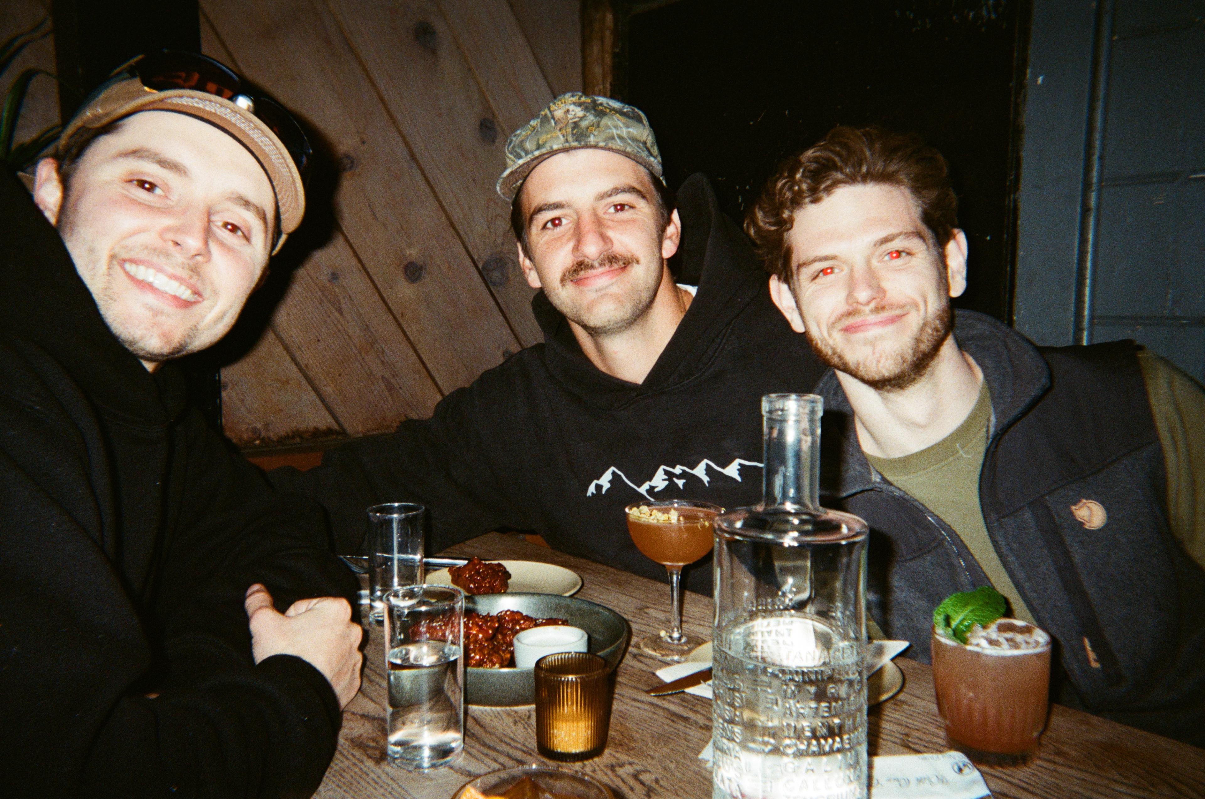 Three smiling men gather around a restaurant table with cocktails, water and shared dishes.